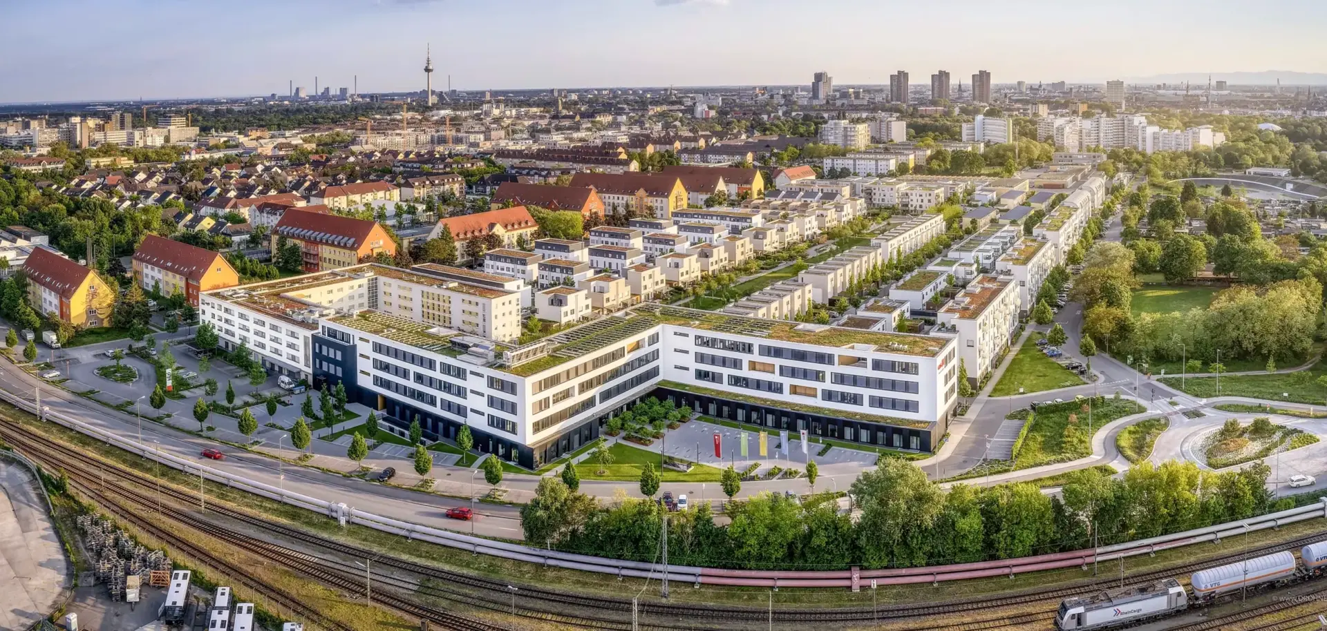 KI-basierte Bildbeschreibung: Luftaufnahme eines modernen Wohn- und Geschäftskomplexes mit begrünten Dächern, umgeben von Bäumen, Straßen und Bahngleisen, mit einer Stadtlandschaft im Hintergrund unter klarem Himmel.