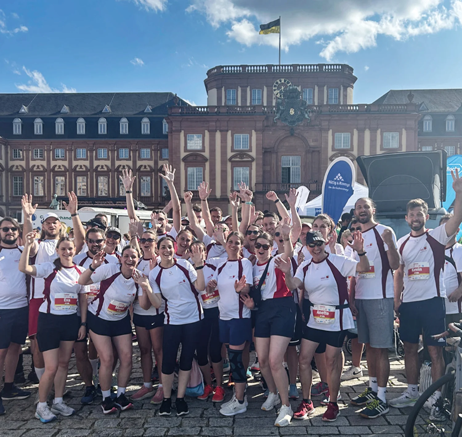 KI-basierte Bildbeschreibung: Eine große Gruppe von Menschen in passenden Sporttrikots lächelt und hebt die Arme. Sie posieren für ein Gruppenfoto im Freien vor einem historischen Gebäude mit einer Uhr und einer Flagge unter einem teilweise bewölkten Himmel.