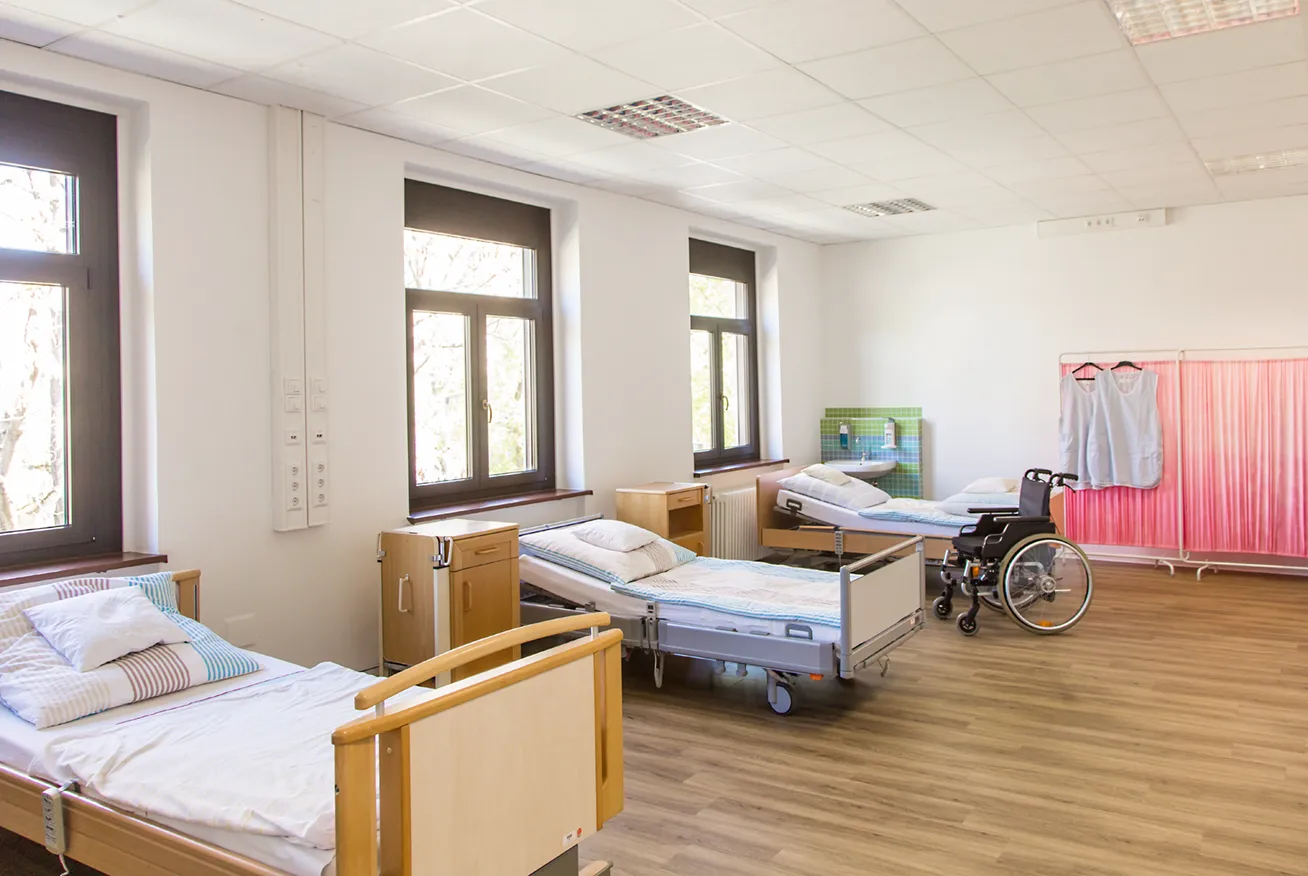 KI-basierte Bildbeschreibung: Ein helles, sauberes Krankenhauszimmer mit drei leeren Betten, einem Rollstuhl, Nachttischen, großen Fenstern und Bademänteln, die an einer Trennwand hängen. Der Raum hat einen Holzfußboden und weiße Wände.