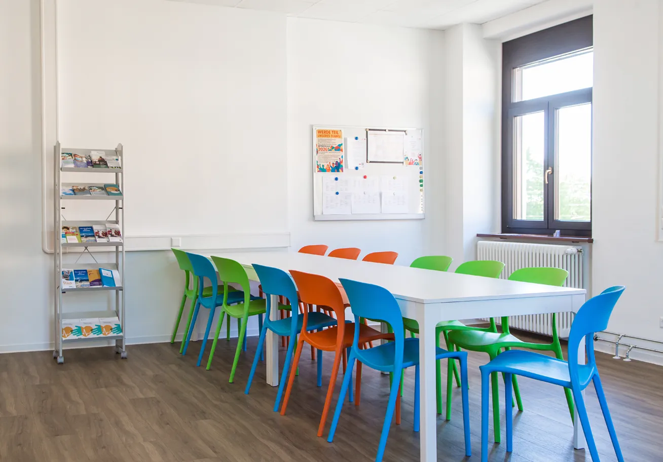 KI-basierte Bildbeschreibung: Ein heller Besprechungsraum mit einem langen weißen Tisch, umgeben von bunten orangefarbenen, grünen und blauen Stühlen, einem Metallregal mit Zeitschriften und einem schwarzen Brett an der Wand neben einem großen Fenster.