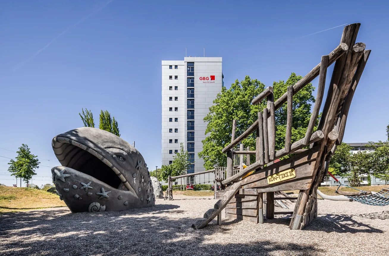 KI-basierte Bildbeschreibung: Ein Spielplatz mit einer großen Walskulptur mit offenem Maul und einem hölzernen Klettergerüst auf einem Kiesplatz mit Bäumen und einem hohen Gebäude im Hintergrund unter einem klaren blauen Himmel.