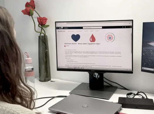 KI-basierte Bildbeschreibung: Eine Person mit langen Haaren sitzt an einem Schreibtisch und blickt auf einen Computermonitor, der eine Website mit Symbolen und deutschem Text anzeigt. Eine Wasserflasche, rote Blumen in einer Vase und ein geschlossener Laptop stehen ebenfalls auf dem weißen Schreibtisch.