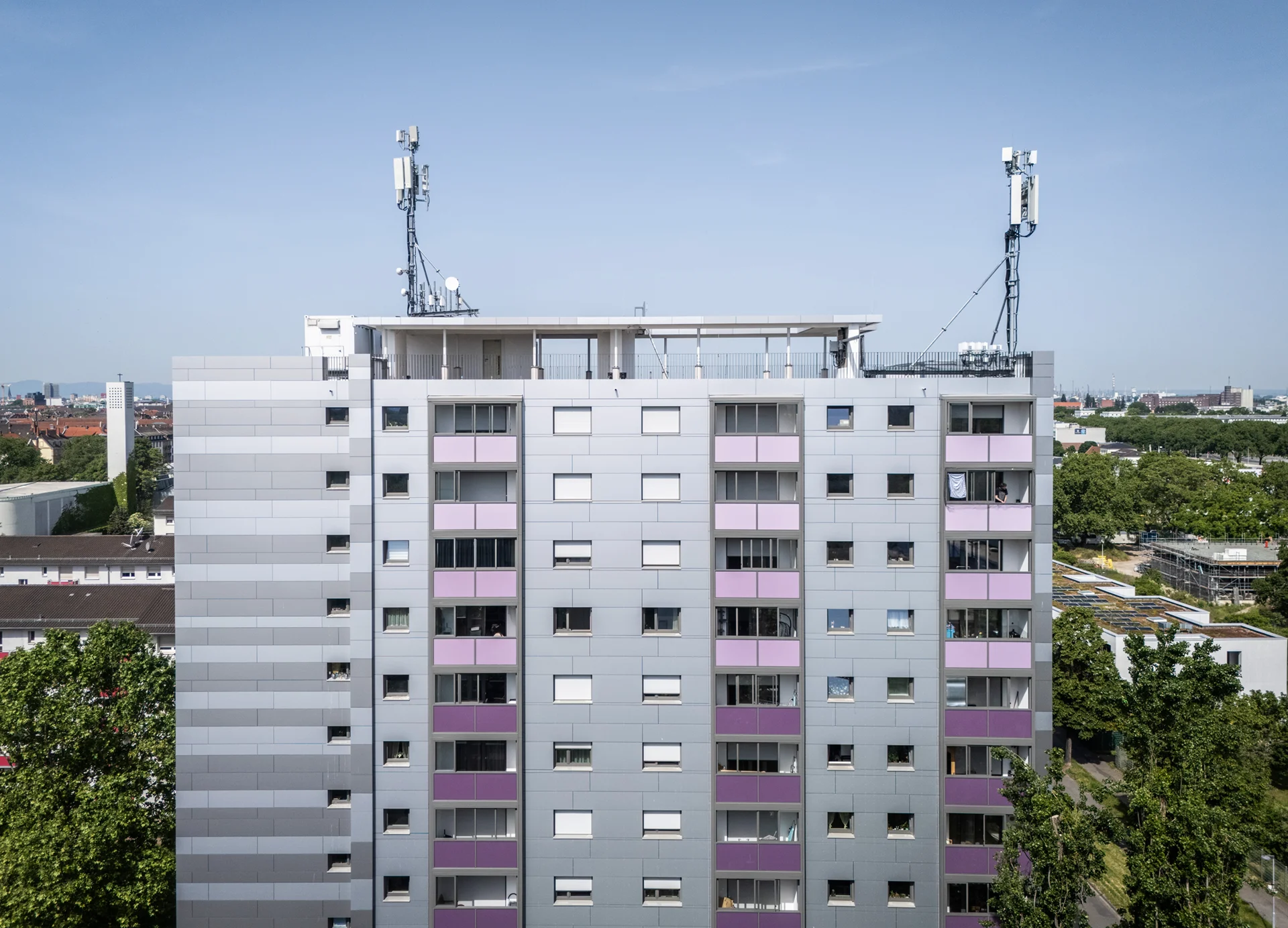 KI-basierte Bildbeschreibung: Ein hohes, modernes Wohngebäude mit grauer und violetter Fassade, zahlreichen Fenstern und zwei großen Zellentürmen auf dem Dach vor einem klaren blauen Himmel und einer Stadtlandschaft im Hintergrund.