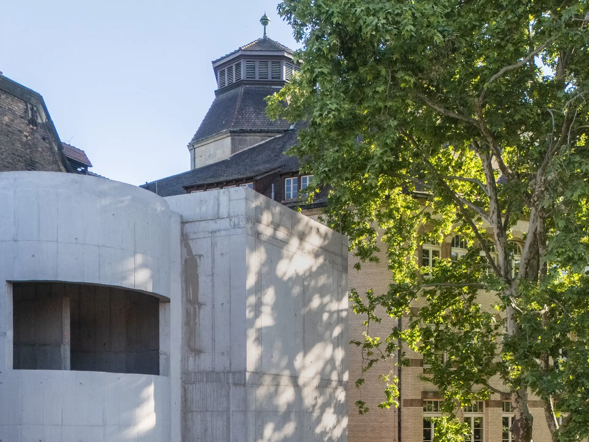 KI-basierte Bildbeschreibung: Ein modernes, im Bau befindliches Betongebäude steht neben einem älteren Backsteingebäude mit gewölbten Fenstern, umgeben von großen, belaubten Bäumen und unter einem strahlend blauen Himmel.