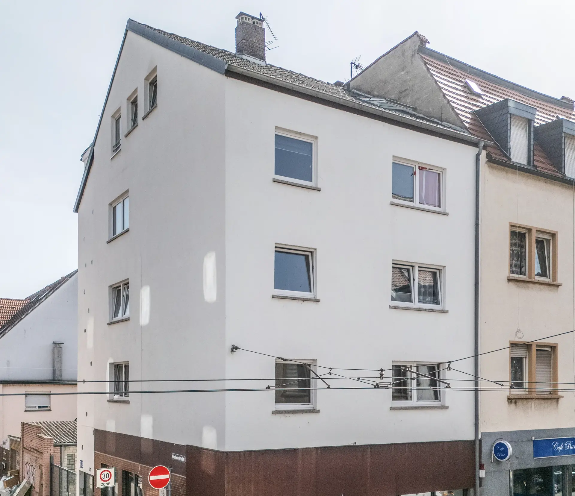 KI-basierte Bildbeschreibung: Ein vierstöckiges Eckgebäude mit weißen Wänden, brauner Verkleidung im Erdgeschoss und mehreren Fenstern. Im Erdgeschoss sind Geschäfte untergebracht, und vor dem Gebäude sind Straßenschilder und Kabelleitungen zu sehen.