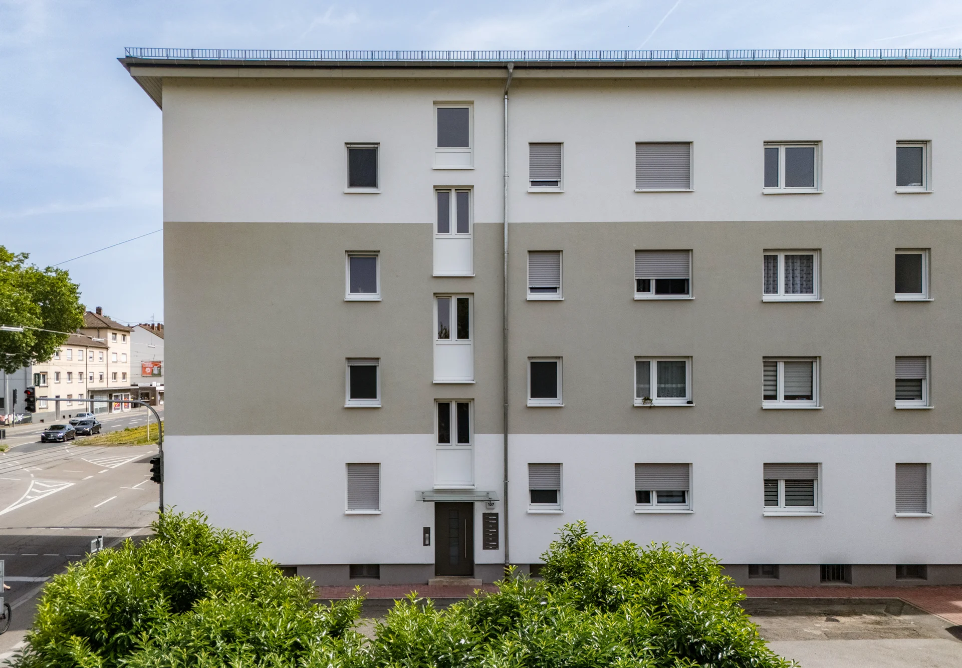 KI-basierte Bildbeschreibung: Ein vierstöckiges Mehrfamilienhaus mit Flachdach, hellgrauer und weißer Fassade, geschlossenen Fensterläden und einem zentralen Eingang; im Vordergrund sind Büsche zu sehen und links ist eine Straße zu erkennen.