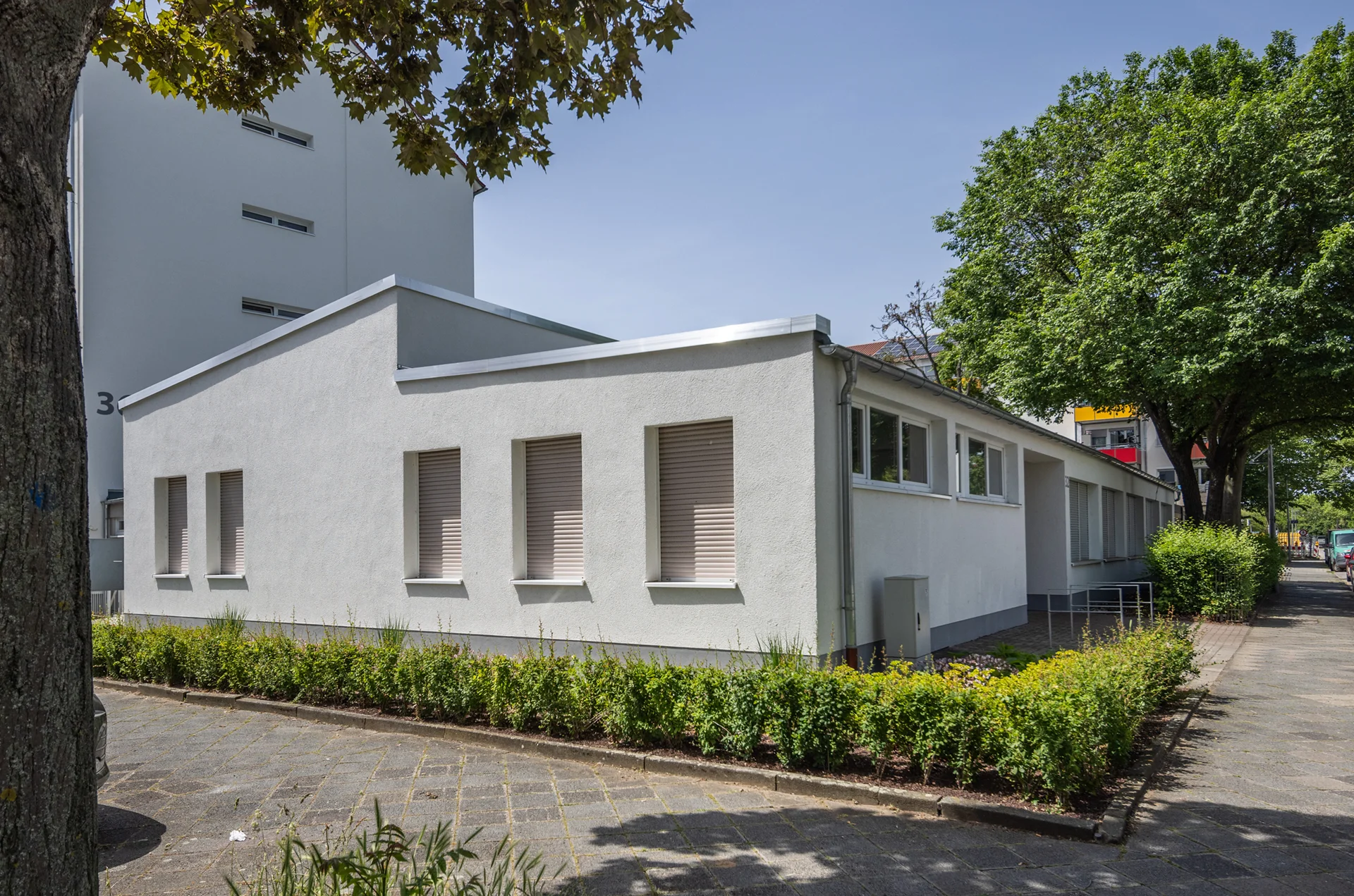 KI-basierte Bildbeschreibung: Ein einstöckiges weißes Gebäude mit geschlossenen Fensterläden, umgeben von grünen Sträuchern und Bäumen, auf einem gepflasterten Bürgersteig unter freiem Himmel.
