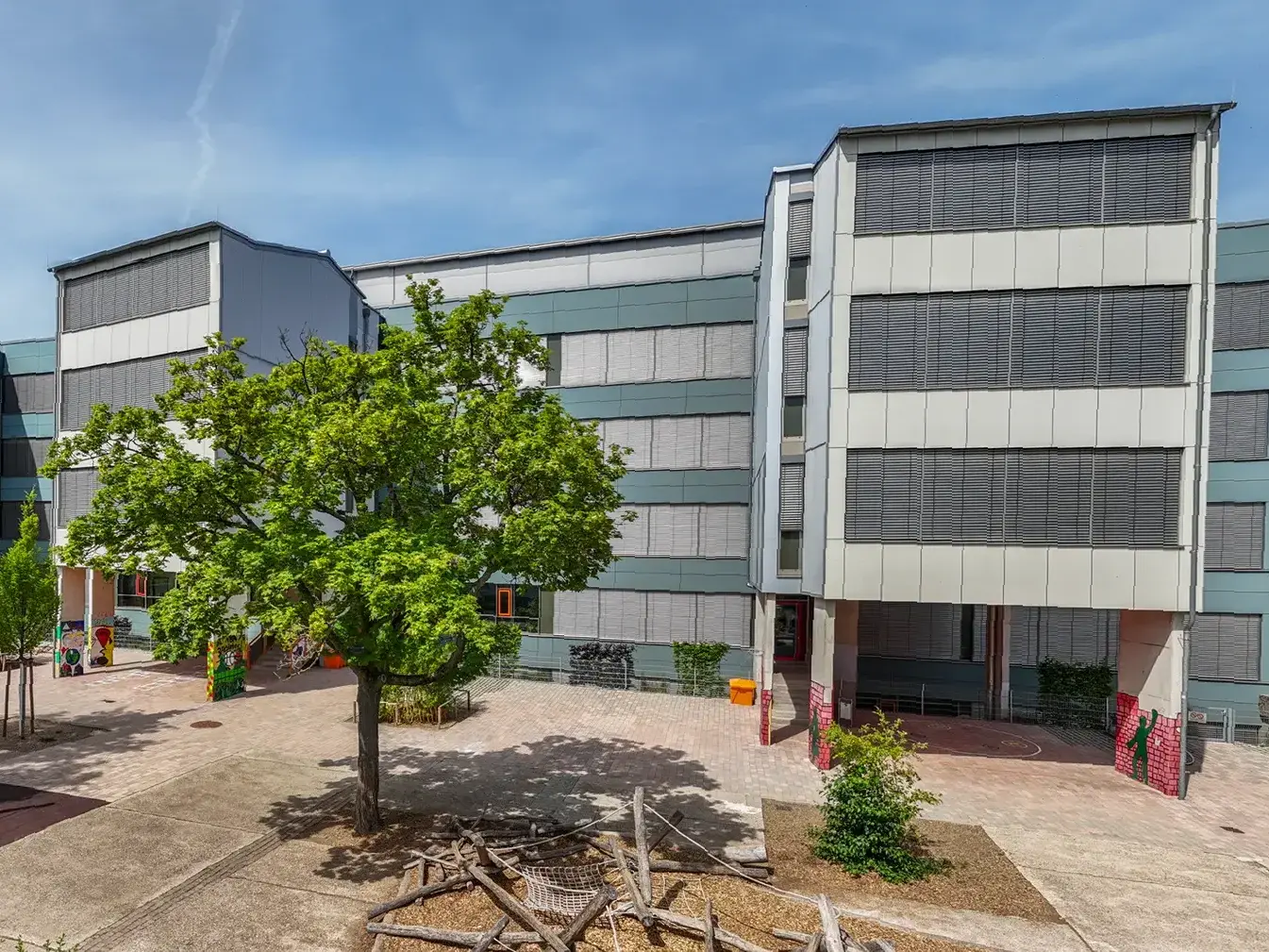 KI-basierte Bildbeschreibung: Ein modernes Schulgebäude mit grauen und grünen Paneelen, großen Fenstern und Bäumen im Innenhof unter blauem Himmel. Auf der Freifläche sind Spielgeräte und Bänke für Kinder zu sehen.