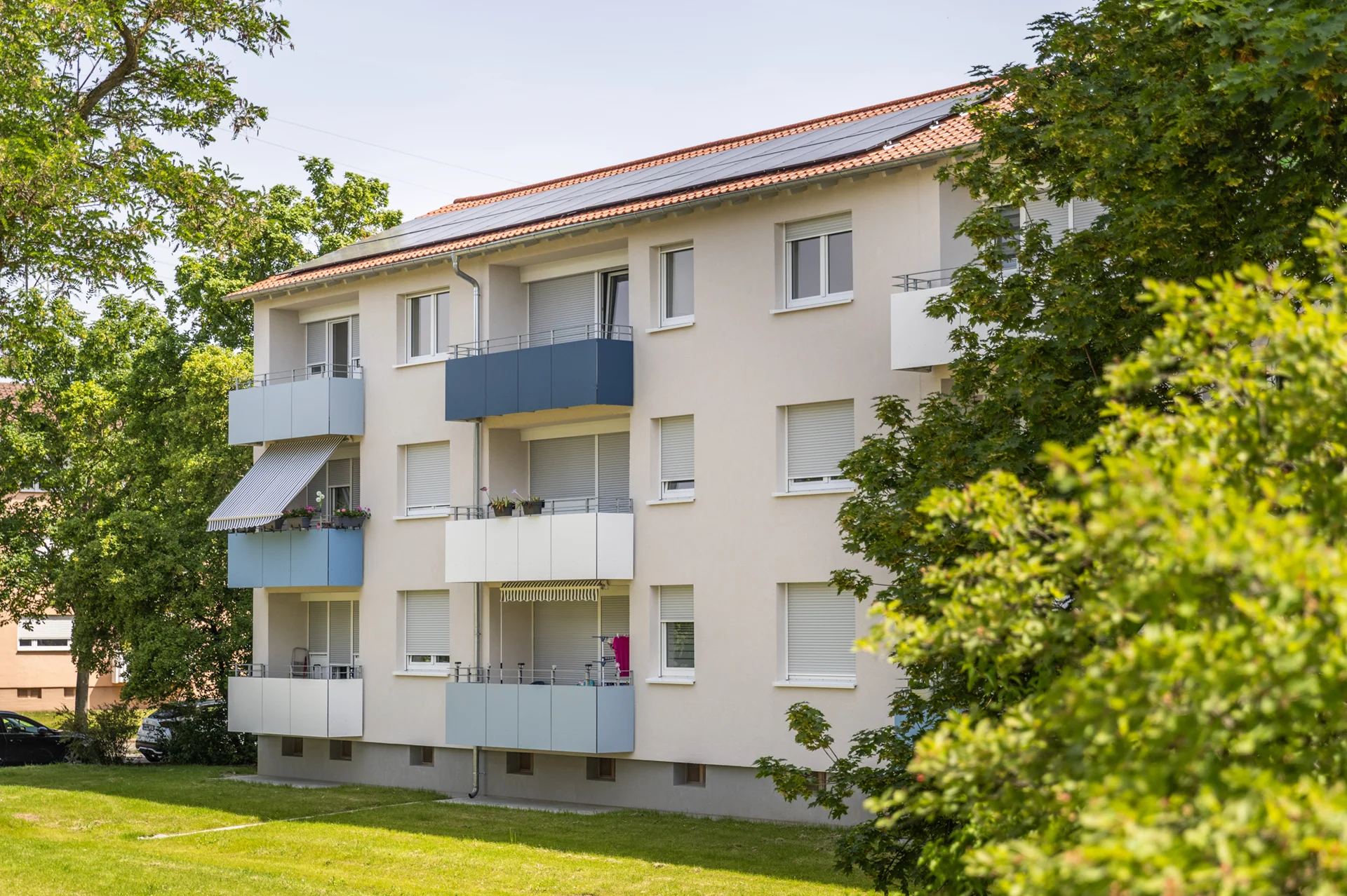 KI-basierte Bildbeschreibung: Ein dreistöckiges Wohnhaus mit Balkonen, weißen Wänden und einem roten Ziegeldach, umgeben von grünen Bäumen und einer Wiese an einem sonnigen Tag.