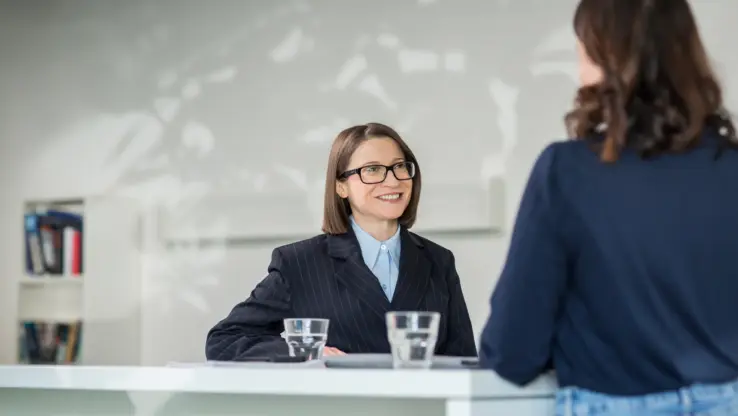 KI-basierte Bildbeschreibung: Zwei Frauen sitzen an einem Schreibtisch und führen ein Gespräch. Eine Frau, die eine Brille und einen dunklen Blazer trägt, lächelt, während sie der anderen zuhört, die mit dem Rücken zur Kamera steht. Auf dem Schreibtisch stehen zwei Gläser mit Wasser.