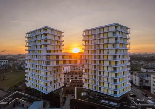KI-basierte Bildbeschreibung: Zwei moderne Hochhäuser mit gelben und weißen Balkonen sind durch eine horizontale Struktur nahe der Spitze verbunden. Im Hintergrund geht die Sonne unter und wirft ein warmes Licht auf die Stadtlandschaft.