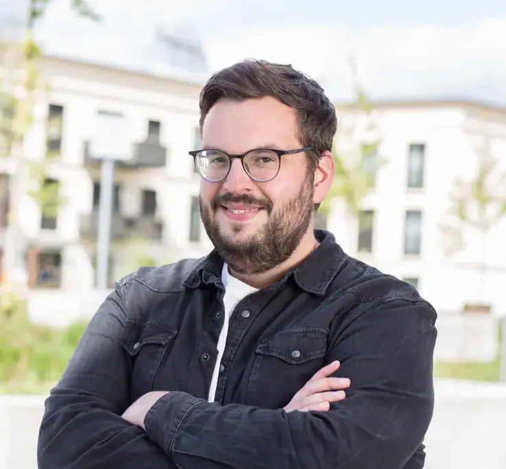 KI-basierte Bildbeschreibung: Ein Mann mit dunklem Haar, Brille und Bart lächelt mit verschränkten Armen. Er trägt eine schwarze Jacke über einem weißen Hemd und steht im Freien, wobei ein modernes Gebäude und Bäume im Hintergrund verschwimmen.
