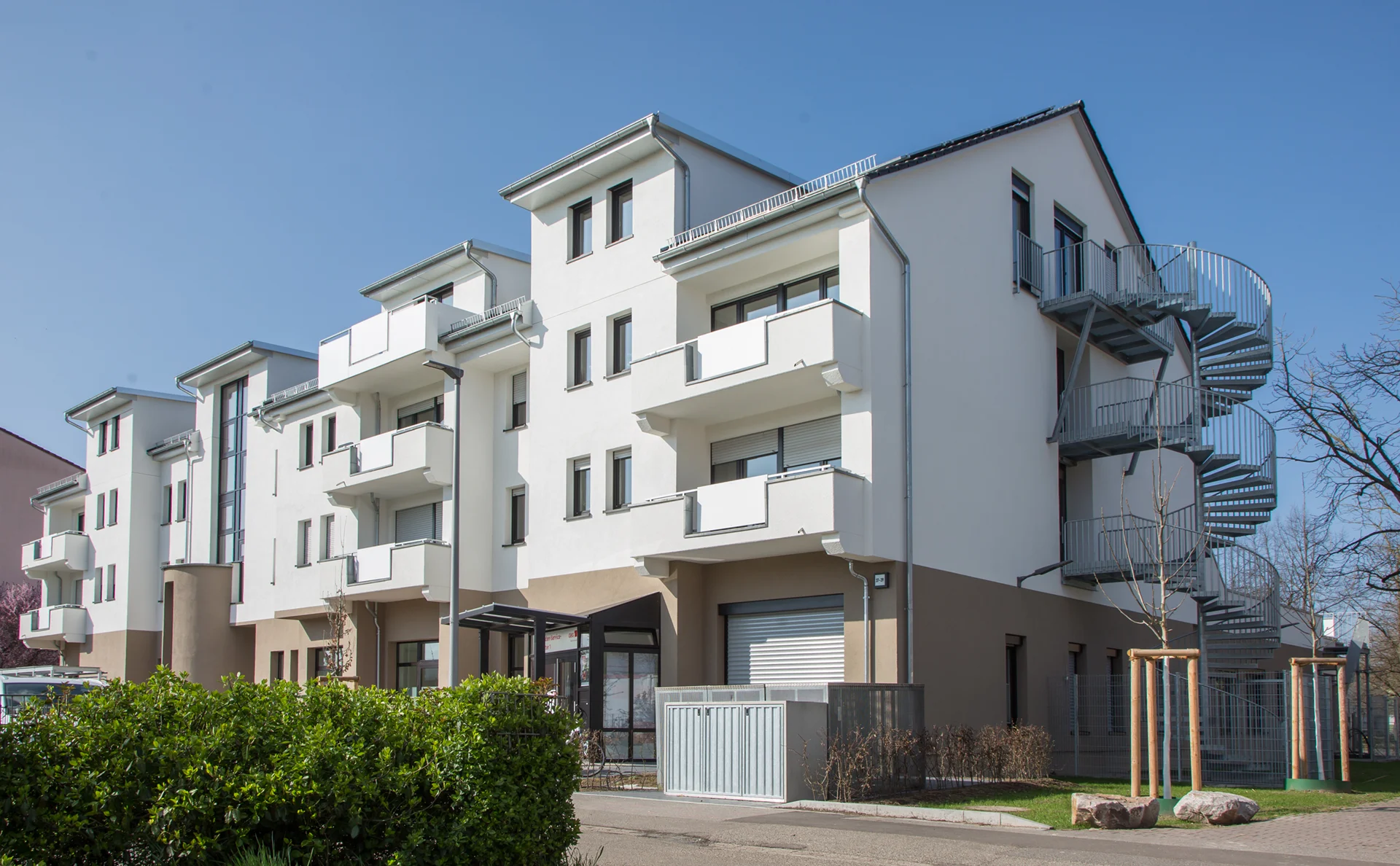 KI-basierte Bildbeschreibung: Ein modernes dreistöckiges Mehrfamilienhaus mit weißen Wänden, mehreren Balkonen und einer externen Wendeltreppe, an einem sonnigen Tag mit blauem Himmel und etwas Grün im Vordergrund.