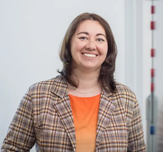 KI-basierte Bildbeschreibung: Eine lächelnde Frau mit schulterlangem braunem Haar, die einen karierten Blazer über einem orangefarbenen Oberteil trägt, steht in einem hellen Innenraum.