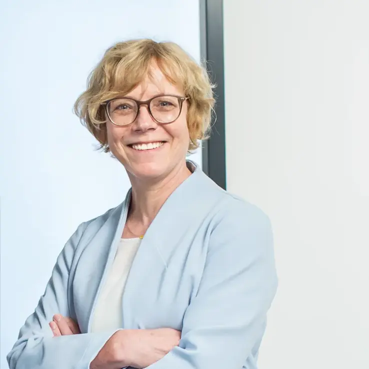 KI-basierte Bildbeschreibung: Eine Frau mit kurzem, lockigem blondem Haar und Brille lächelt selbstbewusst mit verschränkten Armen. Sie trägt einen hellblauen Blazer und steht vor einem hellen, minimalen Hintergrund.