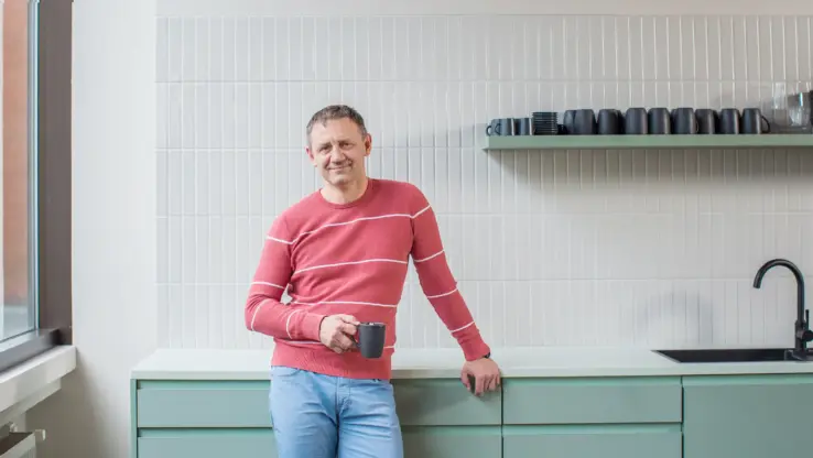 KI-basierte Bildbeschreibung: Ein Mann in einem rot-weiß gestreiften Pullover und einer blauen Hose steht in einer modernen Küche, hält eine schwarze Tasse in der Hand und lächelt. Über einem grünen Tresen mit Spüle befinden sich Regale mit schwarzen Tassen und Schüsseln.