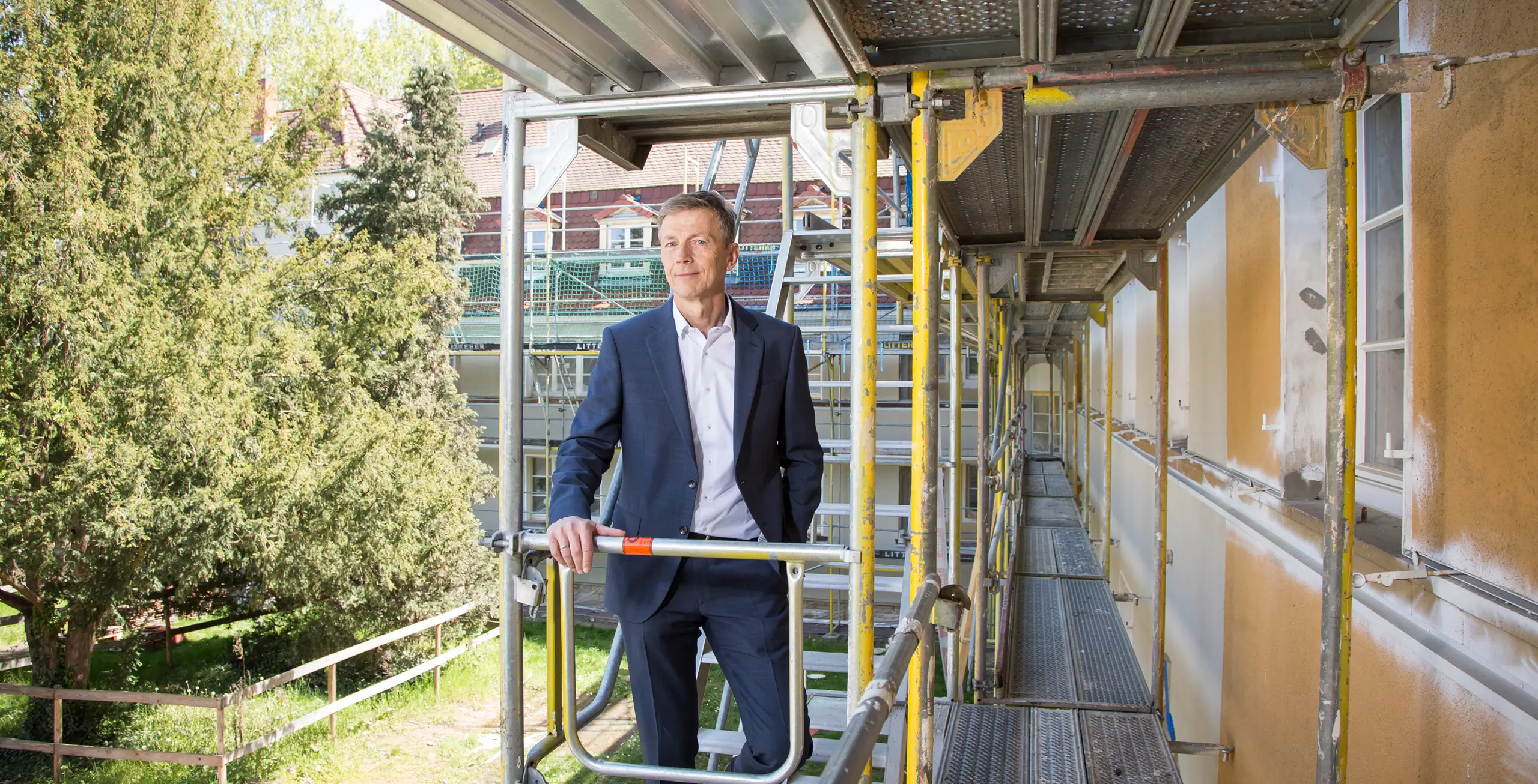 KI-basierte Bildbeschreibung: Ein Mann in einem blauen Anzug steht auf einem Gerüst neben einem Gebäude, das gerade renoviert wird. Im Hintergrund sind an einem sonnigen Tag Bäume und Häuser zu sehen.