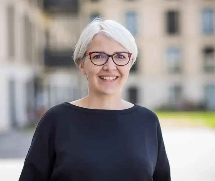 KI-basierte Bildbeschreibung: Eine lächelnde Frau mit kurzen weißen Haaren und rotgerahmter Brille steht draußen und trägt ein schwarzes Oberteil. Im Hintergrund sind unscharfe Gebäude und Grünflächen zu sehen.