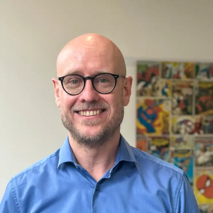 KI-basierte Bildbeschreibung: Ein lächelnder, kahlköpfiger Mann mit Brille und Bart trägt ein blaues Hemd. Im Hintergrund ist auf einer weißen Wand ein farbenfrohes Kunstwerk im Comic-Stil zu sehen, das unscharf wirkt.