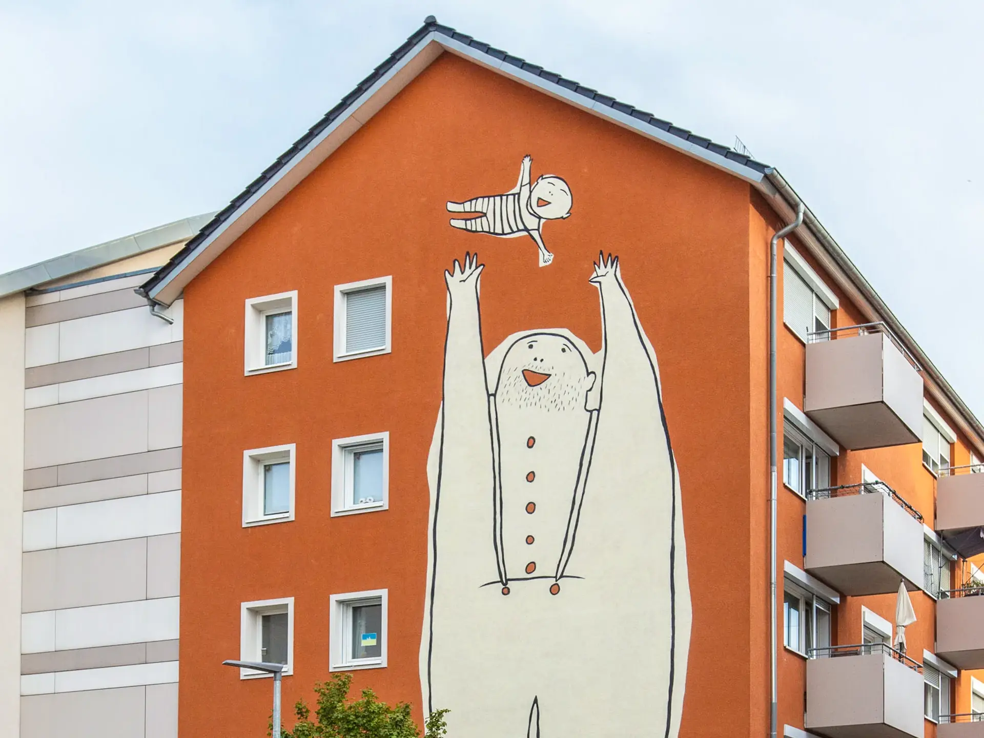 KI-basierte Bildbeschreibung: Ein großes Wandbild an einem orangefarbenen Wohnhaus zeigt eine große, cartoonartige Figur in Weiß, die mit erhobenen Armen eine kleinere Figur über ihrem Kopf hält. Das Wandgemälde zeigt verspielte, einfache Linien und lächelnde Gesichter.