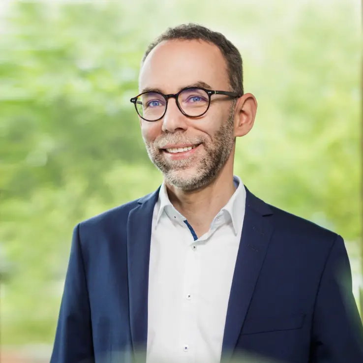 KI-basierte Bildbeschreibung: Ein lächelnder Mann mit kurzen Haaren, Brille und Bart, der ein weißes Hemd und einen dunkelblauen Blazer trägt, steht vor einem verschwommenen grünen Hintergrund im Freien.