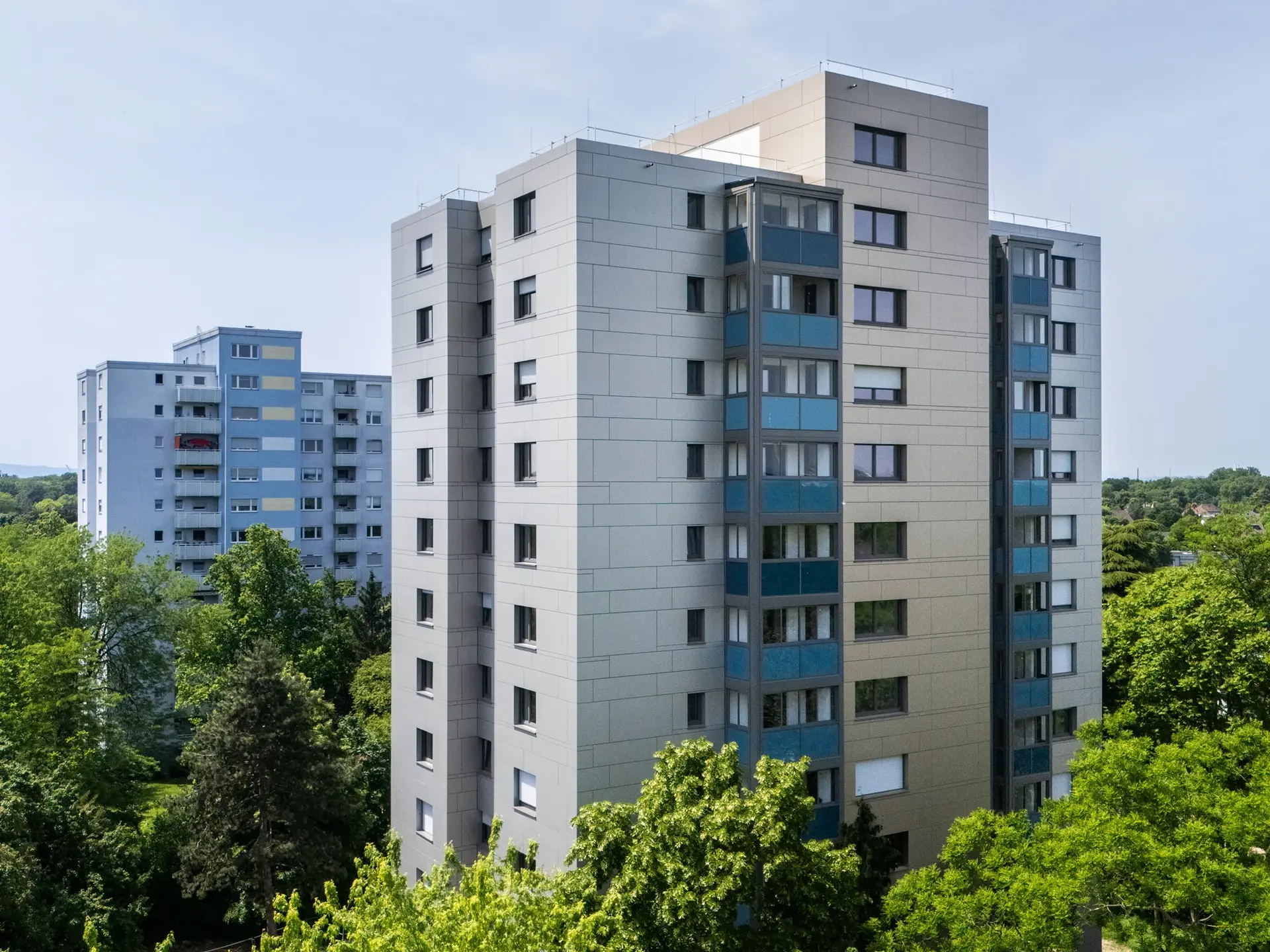 KI-basierte Bildbeschreibung: Zwei moderne, mittelhohe Wohngebäude mit hellen Fassaden und vielen Fenstern, umgeben von grünen Bäumen und Blättern an einem klaren, sonnigen Tag.