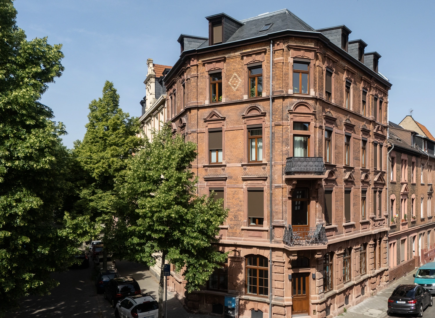 KI-basierte Bildbeschreibung: Ein historisches, vierstöckiges Eckgebäude aus rotem Backstein mit kunstvollen architektonischen Details und einem kleinen Balkon, umgeben von grünen Bäumen und geparkten Autos an einer ruhigen Straße.