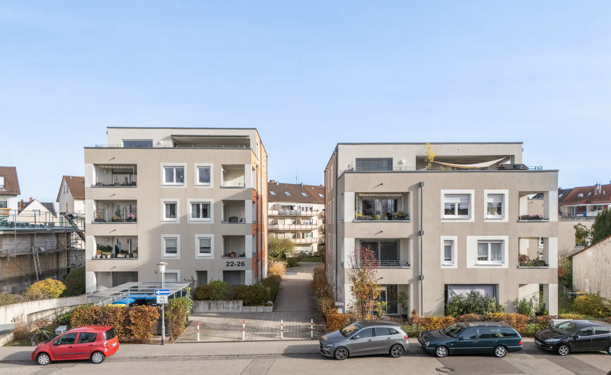 KI-basierte Bildbeschreibung: Zwei moderne, beigefarbene Wohnhäuser mit Balkonen stehen nebeneinander, getrennt durch eine schmale Einfahrt. Vor dem Haus reihen sich geparkte Autos aneinander, und kleine Sträucher und Bäume schmücken die Szenerie unter einem klaren Himmel.