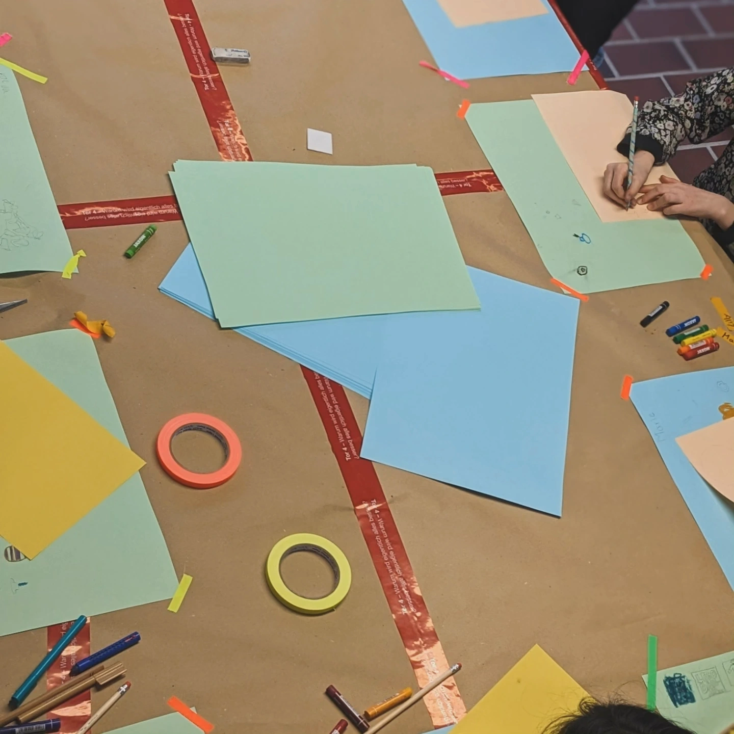 KI-basierte Bildbeschreibung: An einem mit braunem Papier bedeckten Tisch malen und kolorieren Kinder auf pastellfarbenen Blättern. Buntstifte, Farbstifte, Klebeband und anderes Malzubehör liegen verstreut herum. Zu sehen sind nur Hände und Arme.