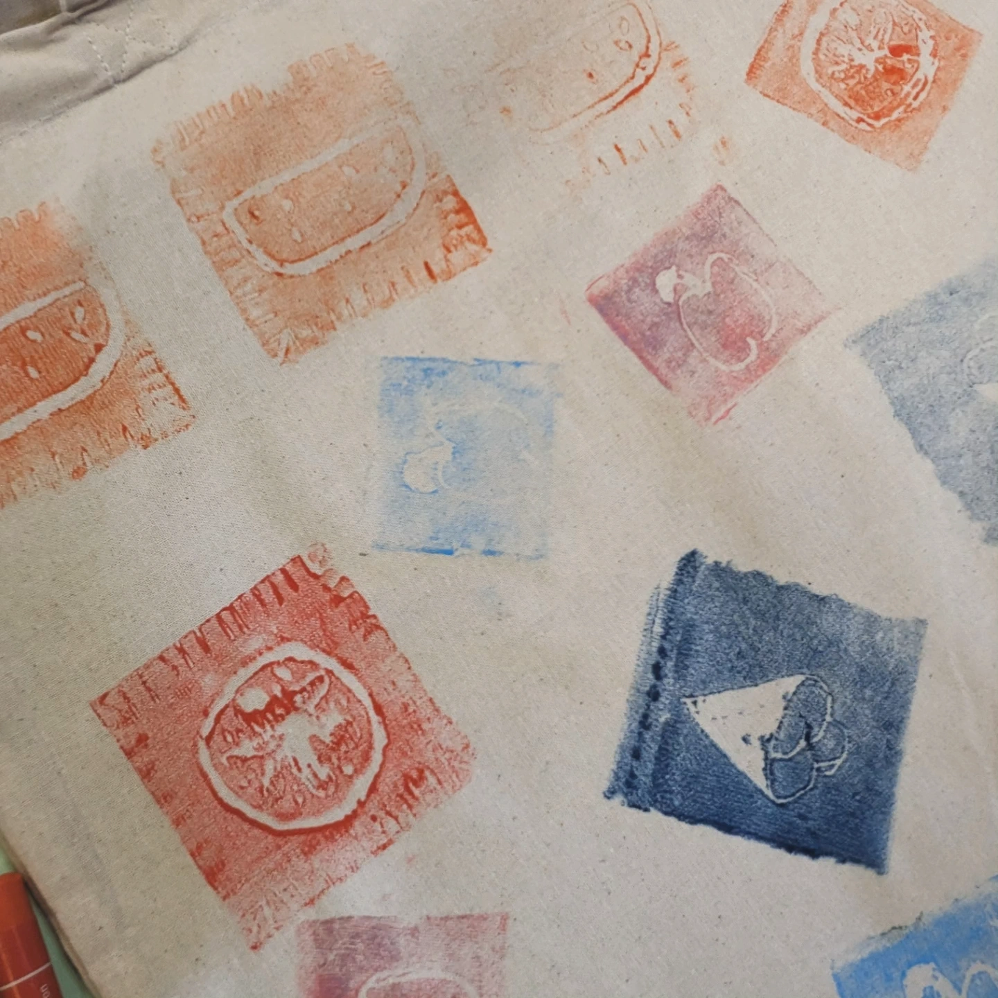 KI-basierte Bildbeschreibung: Ein beiger Stoff ist mit bunten Blockdrucken in verschiedenen Formen und mit Fruchtmotiven, darunter Wassermelonen, sowie weiteren abstrakten Mustern in roter, blauer und rosa Farbe verziert.