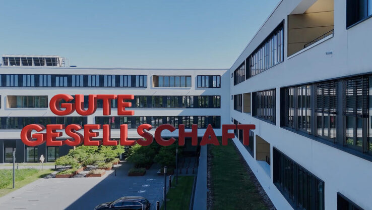 KI-basierte Bildbeschreibung: Moderne Bürogebäude mit großen Fenstern stehen einander gegenüber. Über dem Innenhof, in dem Bäume, Bänke und geparkte Autos zu sehen sind, schwebt ein roter 3D-Schriftzug in deutscher Sprache: „GUTE GESELLSCHAFT“. Der Himmel ist klar und blau.